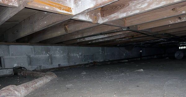 A large metal pipe passes through the shallow dirt crawl space under a home with cinderblock foundation walls.