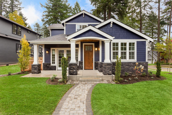 Cute and safe little blue and white house with a green grass lawn backed by very tall and spreading pine trees and a very blue sky with few clouds.